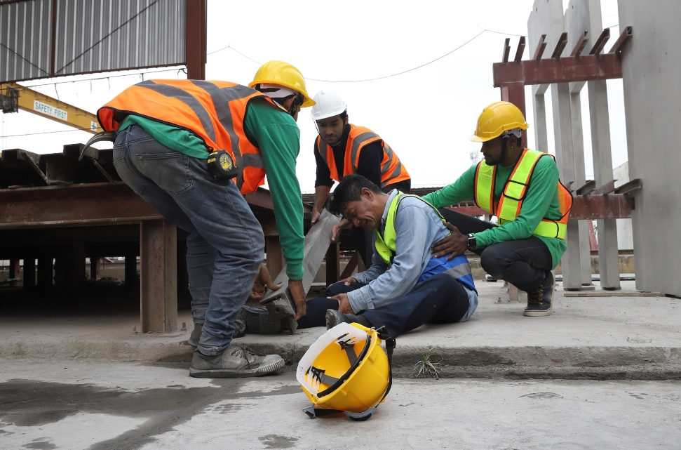 Workers in safety gear assist an injured colleague sitting on the ground. A yellow hard hat lies nearby, a silent witness to the dangerous jobs Latino immigrants undertake. They are on a construction site with concrete structures and beams in the background.