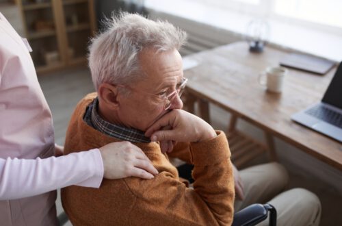 An elderly man in a wheelchair, wearing a brown sweater and glasses, looks thoughtful while resting his chin on his hand. A person in a pink shirt stands beside him with a hand gently placed on his shoulder. Medicare Advantage.