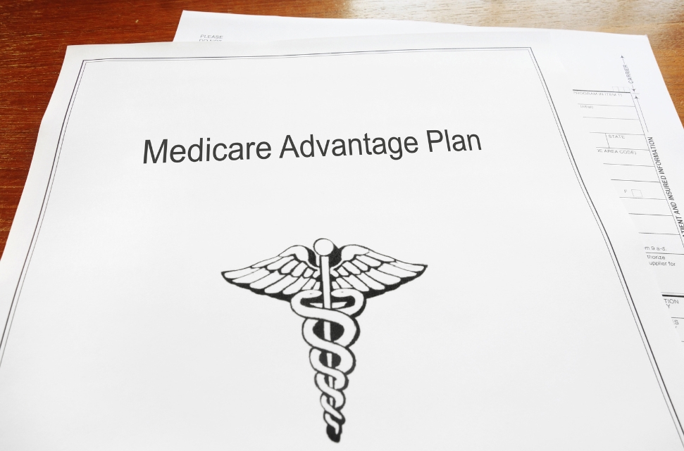 A document titled "Medicare Advantage Plan" lies on a table. Below the title, there is a Caduceus symbol, featuring two snakes wrapped around a winged staff.