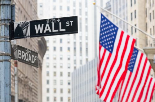 Street signs for Wall Street and Broad Street are visible, with several American flags in the background. Tall buildings line the street, creating an urban atmosphere. Stop Wall Street Looting Act. Private Equity.