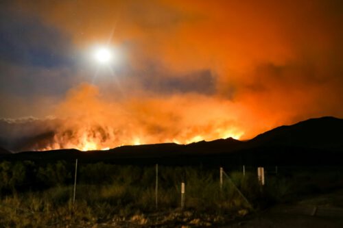 Large wildfires are burning behind mountains, with thick orange smoke billowing into the sky. The glowing sun is visible above the horizon, illuminating the darkened landscape in the foreground increasing concerns about climate change.