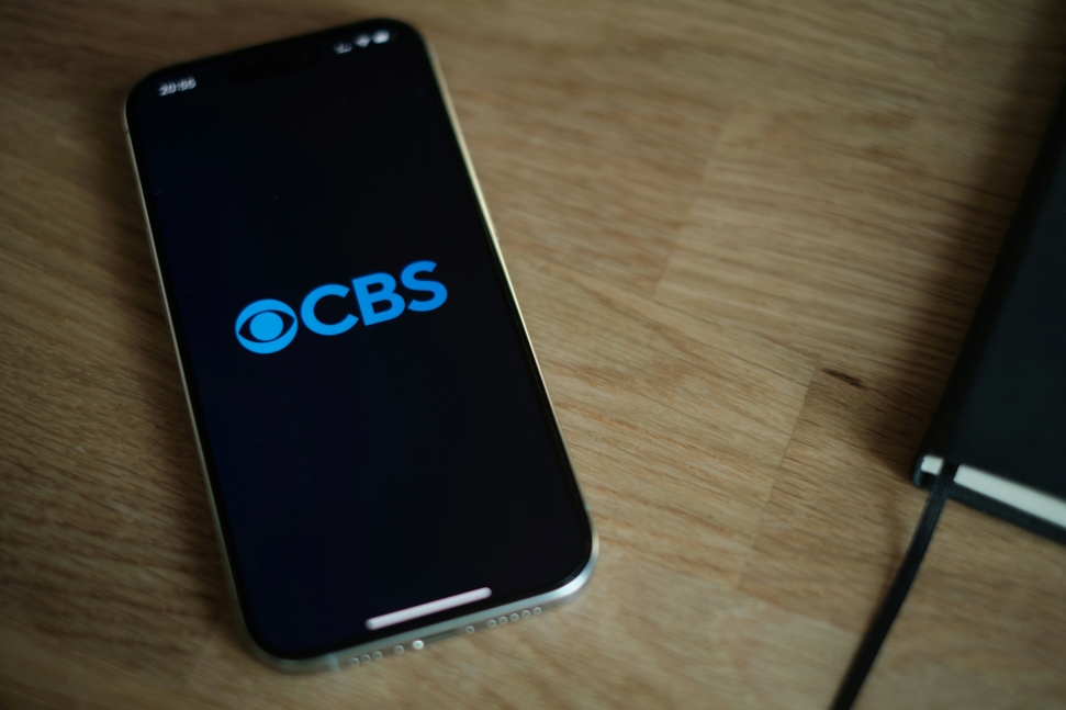 A smartphone with the CBS logo displayed on its screen rests on a wooden surface. A black notebook is partially visible on the right side of the image.