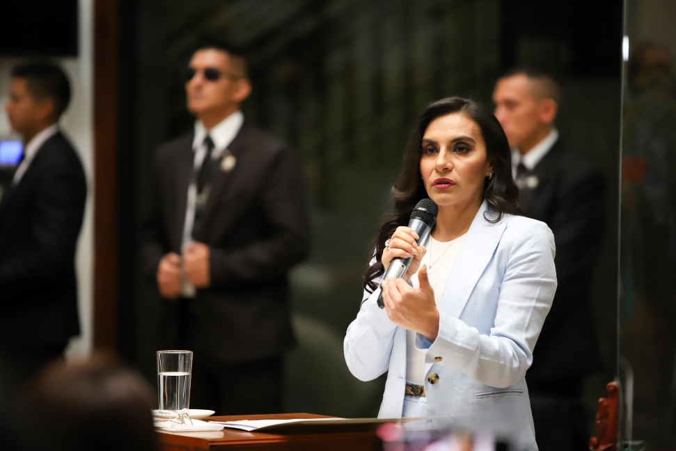 Ecuador News Round Up: Veronica Abad, Vice President of Ecuador, speaks at a press conference. She is in a light blue blazer and speaking into a microphone at a podium. She has long, dark hair and is gesturing with her hand. A glass of water is beside her, and several men in suits are standing in the background.