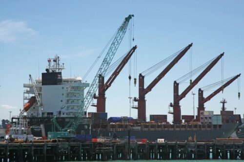 A large cargo ship docked at a port with several cranes unloading containers. The sky is clear, and various containers are stacked on the ship and dock. Photo by Nareeta Martin.