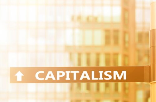 A street sign with the word CAPITALISM and an upward arrow is set against a backdrop of modern glass buildings, suggesting economic growth or progress.