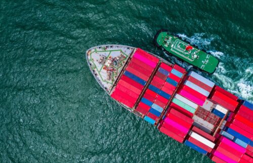 Aerial view of a large container ship with colorful containers on the ocean, accompanied by a small green tugboat.