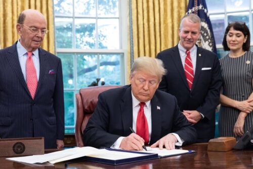 Donald Trump sits at a desk signing papers surrounded by three people: two men in suits and a woman in a striped dress.