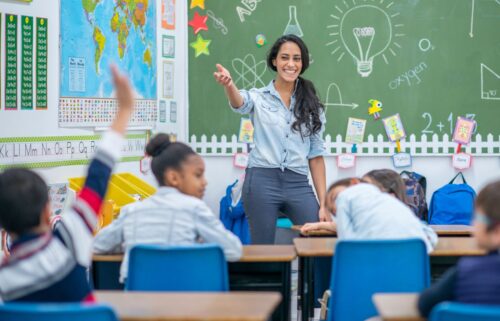 A teacher standing in front of a classroom, engaging with her students who are seated at desks. The classroom is filled with educational materials, and the students are taking notes. The teacher is a woman, dressed professionally, and is actively teaching while pointing to text on the board.