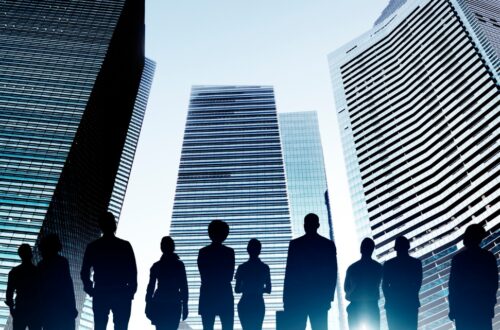 Silhouettes of people stand against a backdrop of tall skyscrapers, where modern glass exteriors reflect a clear blue sky.