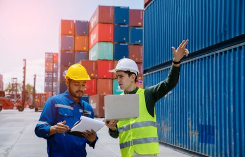 Two workers in safety gear, one with a yellow hard hat and the other with a white hard hat and lime vest, discuss documents. They stand in a shipping yard with stacked cargo containers. One uses a laptop and gestures upwards representing the strong GDP jump in durable goods.