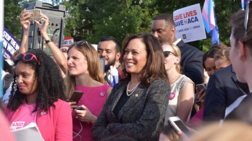 A group of people gathered outdoors to see Vice President Kamala Harris at a protest march. Many are wearing pink, smiling and holding phones. Some hold signs in support of the ACA, reading Save Lives Save the ACA. Trees and a bit of sky are visible in the background.