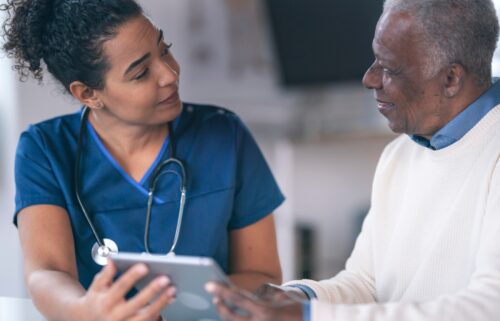 A doctor is engaged in conversation with a patient in an indoor setting. The doctor appears attentive, showing concern for the patient's well-being. Both individuals are focused on each other; the patient is listening intently to possible upcoming cuts to Medicaid.