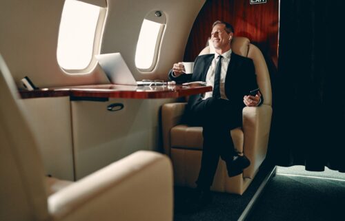 A male corporate CEO sitting on a private jet airplane, with a wall, windows and mirror visible in the background.