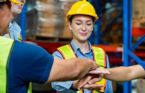 A group of warehouse workers in safety vests and helmets stack their hands in a team gesture, embodying teamwork and cooperation amid an industrial setting of shelves and boxes. Despite the backdrop of union membership decline, their unity shines through.