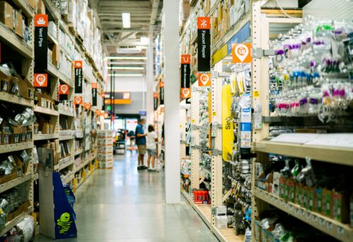 A wide aisle in a hardware store is seen, filled with shelves of various home improvement items. Signs hang above, indicating different product sections. Two people are visible in the distance, browsing. The floor is clean, with bright overhead lighting.