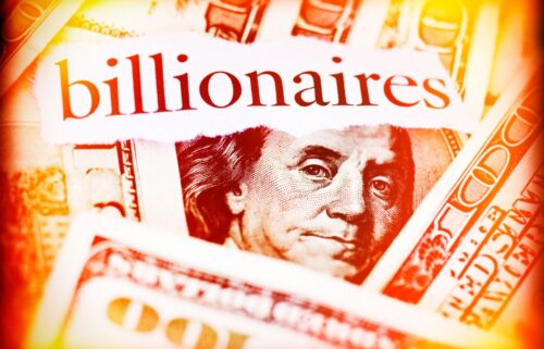 Close-up of scattered 100-dollar bills with Benjamin Franklins portrait visible. The word billionaires is prominently displayed in red on a torn piece of paper placed over the bills, with a warm, glowing effect around the edges.