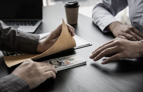 Two people sit at a table whilet he one on the left discreetly passes a bribe to the other. A stack og one hundred dollar billa is visible on a grey table. A coffee cup is in the background.