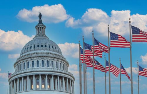 The U.S. Capitol building with its dome is on the left against a blue sky with white clouds. Multiple American flags wave on poles to the right.