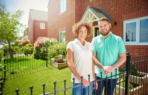 A man and woman stand outdoors in front of a wooden fence and a house, smiling. They are surrounded by grass and plants, with trees in the background.