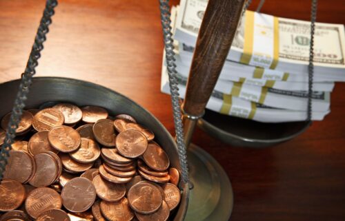 Close-up of a balancing scale with a shallow pan full of pennies on the left and stacks of U.S. hundred-dollar bills on the right. The scale is set against a wooden surface, symbolizing the contrast between small change and large sums of money.