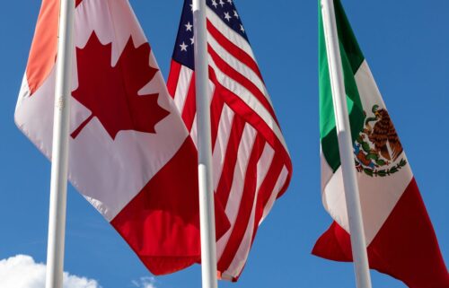 Flags of Canada, the United States, and Mexico waving against a clear blue sky. The Canadian flag features a red maple leaf, the U.S. flag has red and white stripes with stars, and the Mexican flag shows an eagle with a snake on a cactus.