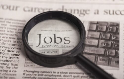 A magnifying glass focuses on the word Jobs in a newspapers employment section. The background shows a part of a keyboard and blurred text.