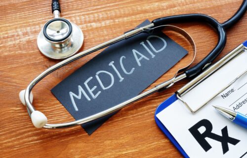 A stethoscope rests on a wooden surface next to a black card with MEDICAID written in white. Beside it, there's a prescription pad with a blue clipboard and pen.