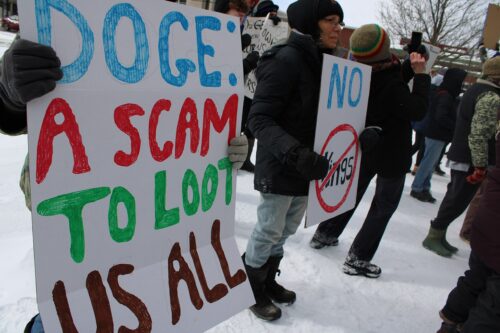 A group of people standing outdoors in winter clothing, holding signs with handwritten messages. The signs contain phrases such as 