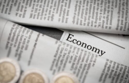 Close-up of a newspaper with the headline Economy prominently displayed. The paper is partially folded, with blurred text surrounding the headline. In the foreground, there are out-of-focus coins. The image suggests financial news or economic reporting.