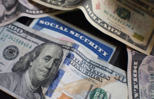 A close-up of scattered U.S. currency and a blue Social Security card. Prominently featured is a $100 bill with Benjamin Franklins portrait, a partially visible $50 bill above, and a corner of a $20 bill in the background. The black surface underneath highlights the money.