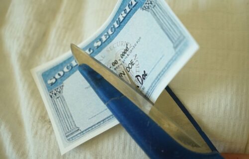 Close-up of scissors cutting a Social Security card on a beige fabric surface. The card features a blue border with white text, partially revealing the words Social Security at the top. The scissor blades are metallic, with blue handles, poised to slice through the card diagonally.