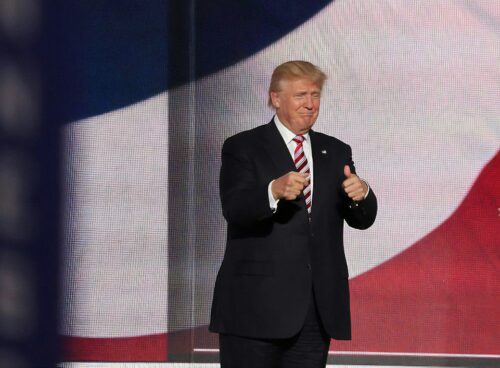 Donald Trump giving a thumbs up in front of an American flag being projected on a large screen.