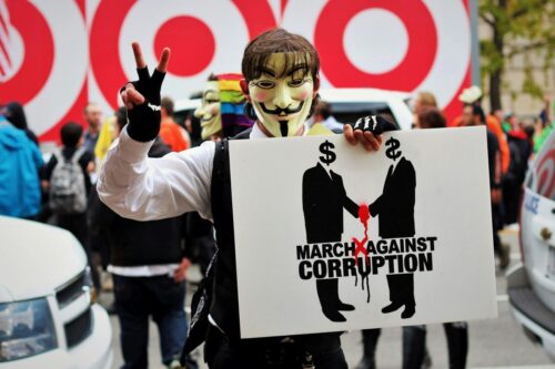 A person wearing a Guy Fawkes mask and a rainbow scarf flashes a peace sign while holding a protest sign that reads March Against Corruption with an image of two figures shaking hands, one with a dollar sign and the other with a rubber glove, amid a crowd and Target signs. Photo by Vincent M.A. Janssen: https://www.pexels.com/photo/photo-of-person-wearing-guy-fawkes-mask-2698473/