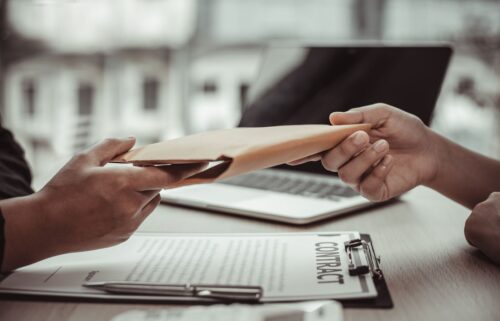 Two people exchanging a brown envelope over a desk. One hand extends the envelope while the other receives it. On the desk are a contract with visible but unreadable text, a pen, and a laptop in the background.