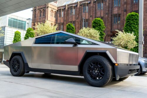 Cybertruck parked on a street. Large, black wheels accent the streamlined design. Background includes a historic brick building with arched windows, green vines, and blooming trees; a modern glass structure is partially visible. Photo by Mylo Kaye on Unsplash