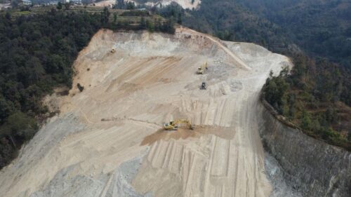 Aerial view of a large construction site on a hillside, with multiple excavators evenly spaced across the cleared earth. Terraced sections and forested areas are visible in the background. The sky is overcast, suggesting a subdued atmosphere. Photo by Raju Shrestha: https://www.pexels.com/photo/construction-work-in-progress-20434886/