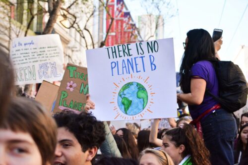 A crowd of people is gathered at a climate protest. A prominent sign in the center reads There is no Planet B with an illustration of Earth. The attendees hold various banners advocating for environmental action. The background shows trees and buildings on a sunny day. Photo by Li-An Lim on Unsplash