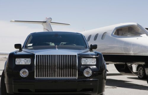 A black luxury car with a prominent chrome grille is parked on an airport runway. Behind it, a sleek private jet is positioned, showcasing its tail and fuselage. The sky is clear and blue, suggesting a bright day.