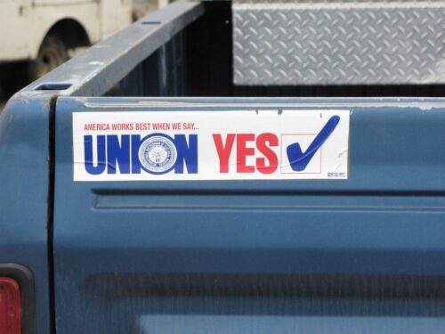 A sticker on a blue truck reads America Works Best When We Say... UNION YES in bold red and blue letters. A checkmark inside a blue box appears to the right. The background includes part of a textured metal surface. Photo: https://www.flickr.com/photos/hartnupj/2739310807/