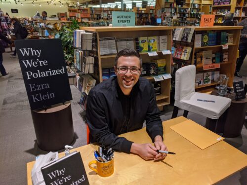 Ezra Klein at a wood desk on a book tour promoting his book 