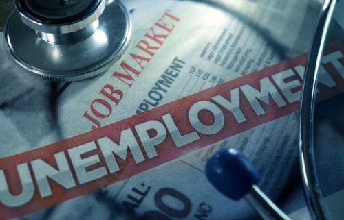 Close-up image of a stethoscope placed on top of a newspaper. The headline UNEMPLOYMENT is prominently displayed in bold red and white letters. Above it, the words JOB MARKET and parts of an unemployment report are visible, suggesting a focus on economic and health issues.