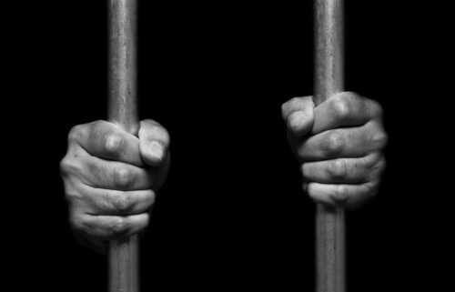 Close-up of two hands tightly gripping vertical metal bars against a black background. The image is in black and white, emphasizing the textures of the hands and bars, suggesting confinement or incarceration.