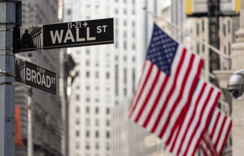 Street signs for Wall Street and Broad Street in focus, with blurred American flags and towering buildings in the background, conveying a financial district atmosphere.