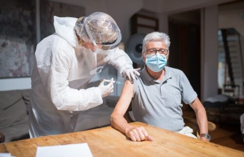 A healthcare worker in protective gear administers a COVID booster vaccine to an elderly man, both wearing masks. The setting is a bright, sunlit room.