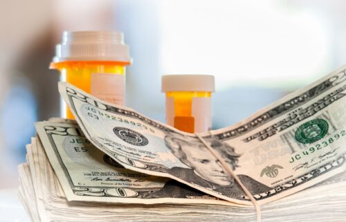 A stack of twenty-dollar bills secured with a rubber band lies in front of orange prescription pill bottles, conveying the cost of medication.