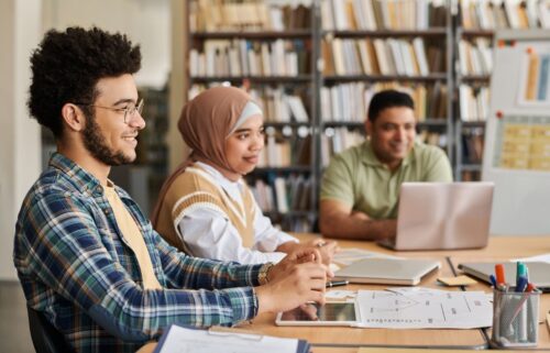 Three foreign students sit at a library table, engaged in discussion. Two use laptops and one holds a tablet. Bookshelves fill the background, suggesting a collaborative and focused atmosphere.