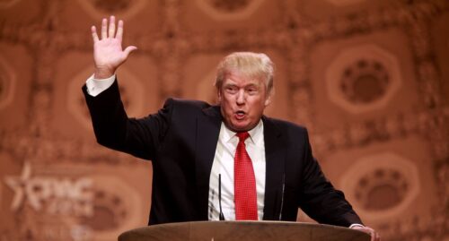 Donald Trump in a dark suit and red tie speaks at a podium, raising his left hand. The backdrop is ornate.