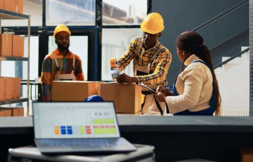Three workers in hard hats and aprons, organize boxes in a warehouse. A laptop in the foreground displays inventory software.