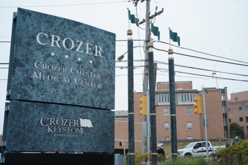 Sign for Crozer-Chester Medical Center in front of a brick building. The image includes green poles with flags, traffic lights, and power lines.