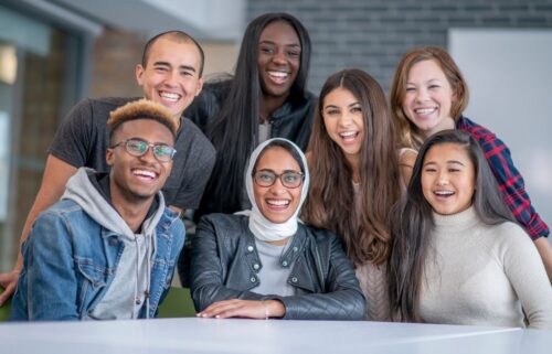 A diverse group of seven people smiling joyfully at the camera. They are casually dressed and appear in a light, spacious indoor setting, conveying warmth and camaraderie.